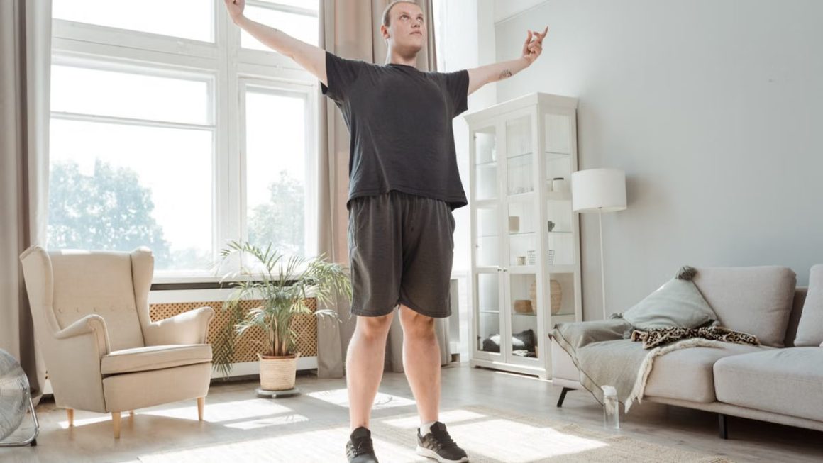 A man performs a stretching exercise with arms out in a bright, modern living room.